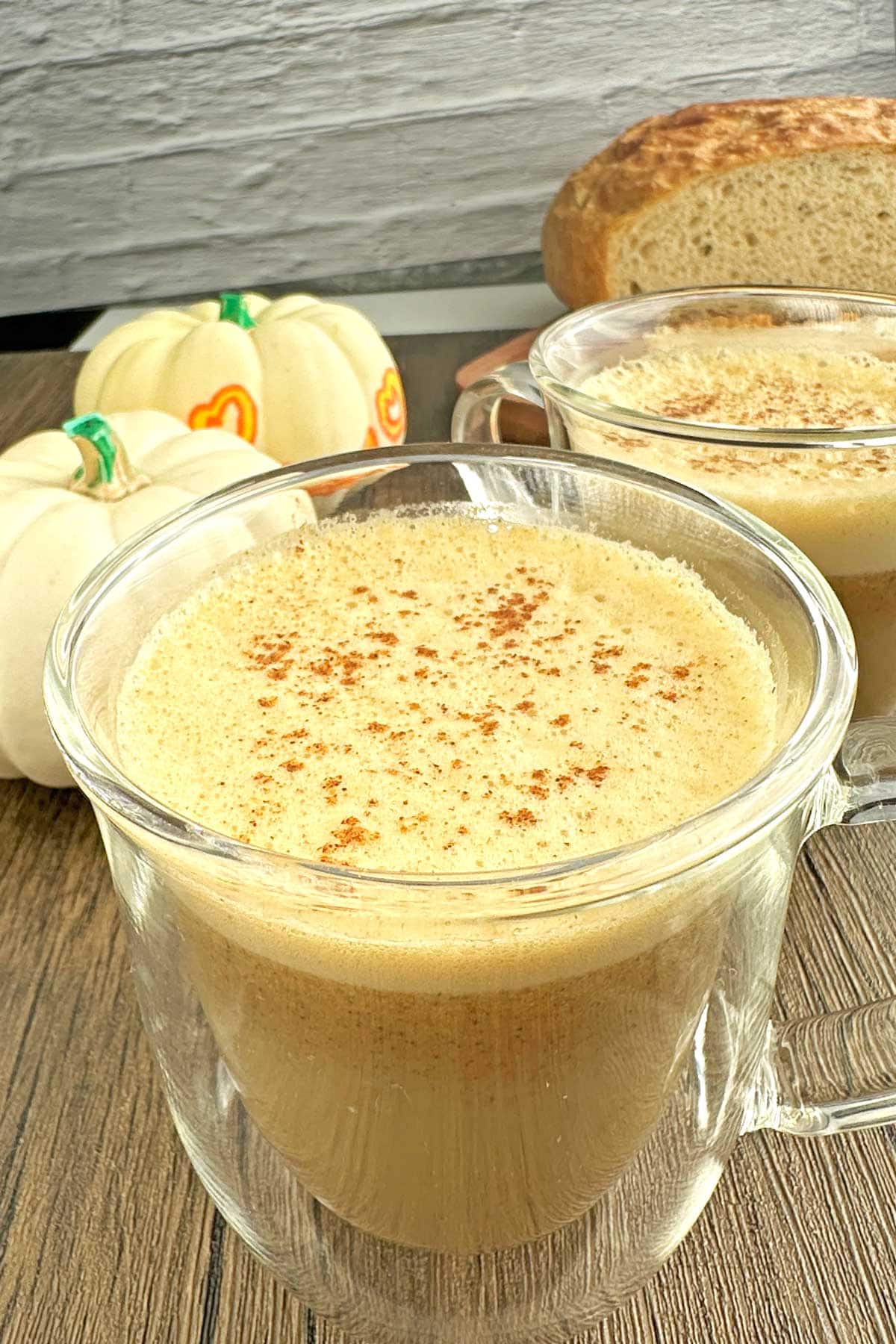 Creamy pumpkin spice latte in glass mugs with cinnamon topping and fresh bread in the background.