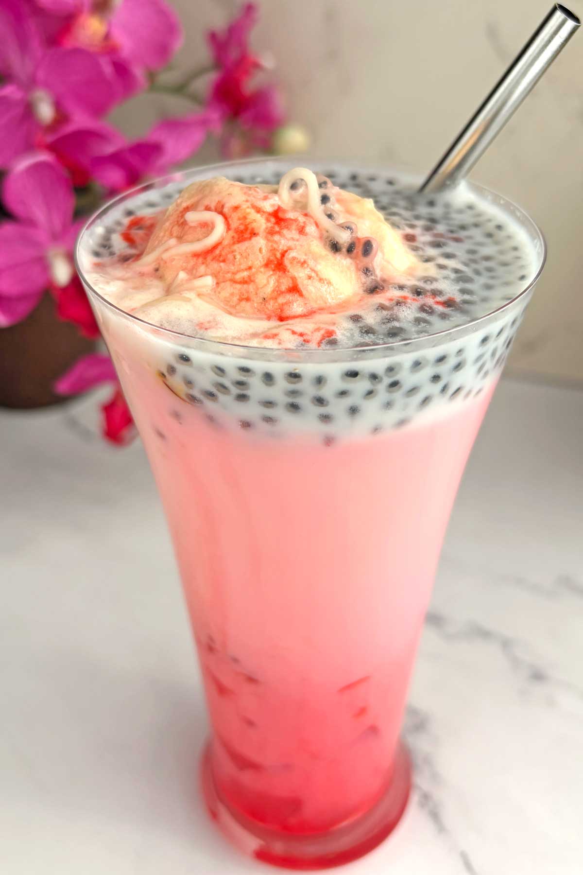 Tall pink bubble tea with black tapioca pearls, topped with a scoop of pale pink ice cream and a metal straw in a clear glass.